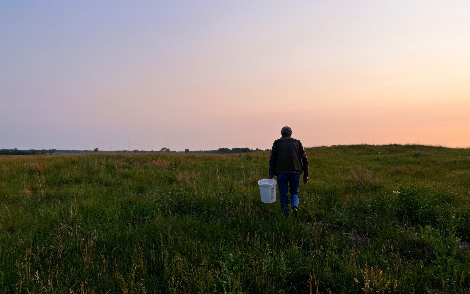 man walking through a field holding a bucket at sunrise