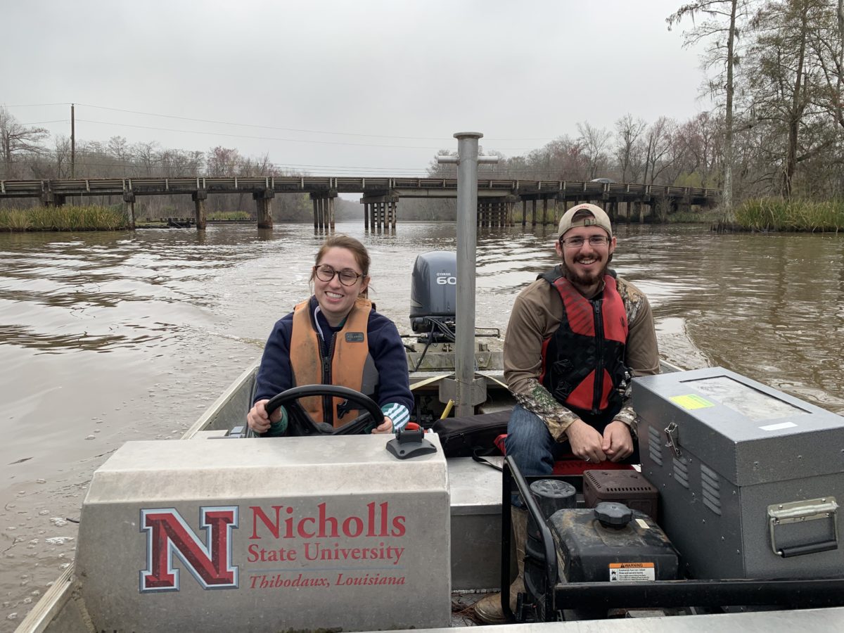 A man and a woman wearing life preservers driving a boat on a brown-water river in Louisiana