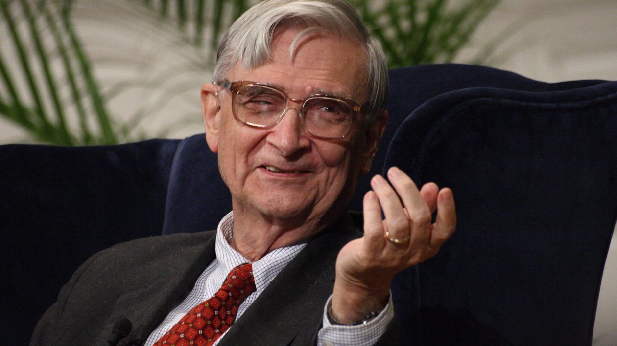 elderly man in a suit and red tie sitting in a chair gesturing