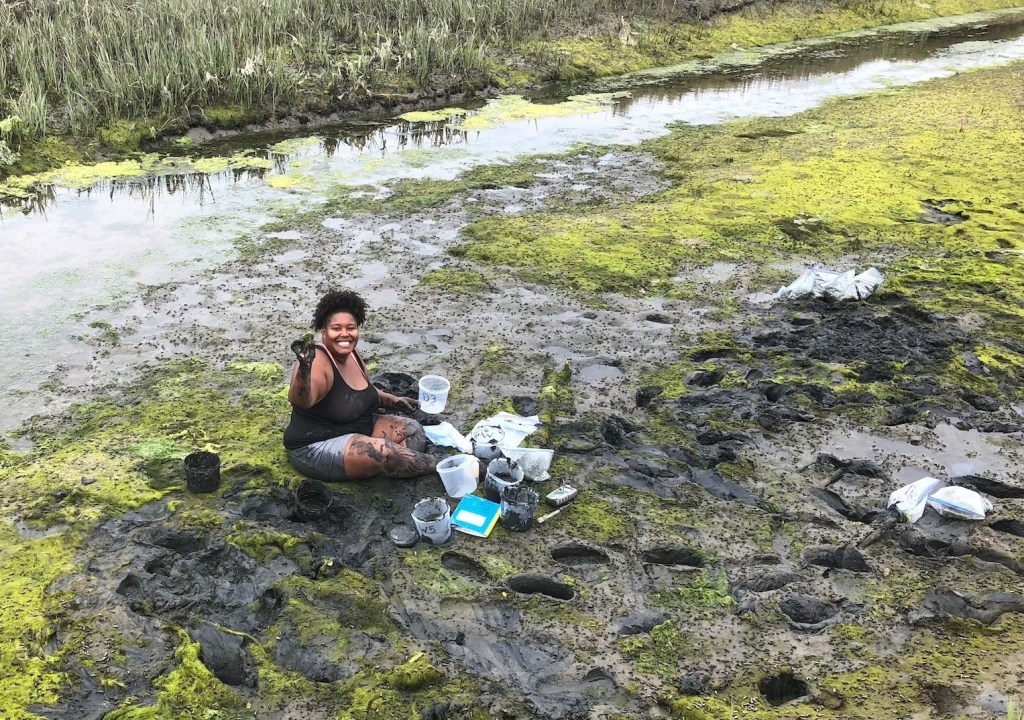 woman sitting in a muddy swamp surrounded by small plastic containers