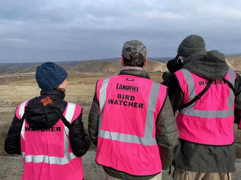 three people in neon pink vests that read BIRDWATCHER looking at a dump