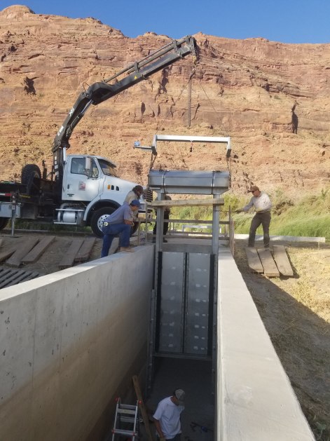 a truck and crane lower a large gate into a concrete passageway. In the background are red sandstone cliffs