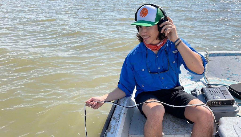 man in a blue shirt, wearing headphones and sitting on a boat holding a microphone overboard