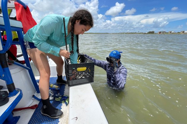 man standing in the water alongside a woman on a boat holding a milk crate