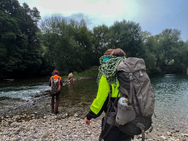 Three people with backpacks on ford a river. The river bottom is covered in stones and there are trees in the background. 