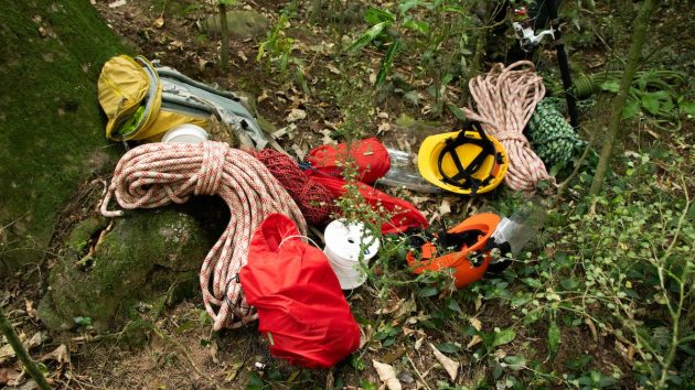 A pile or ropes, helmets, and bags on the forest floor. 