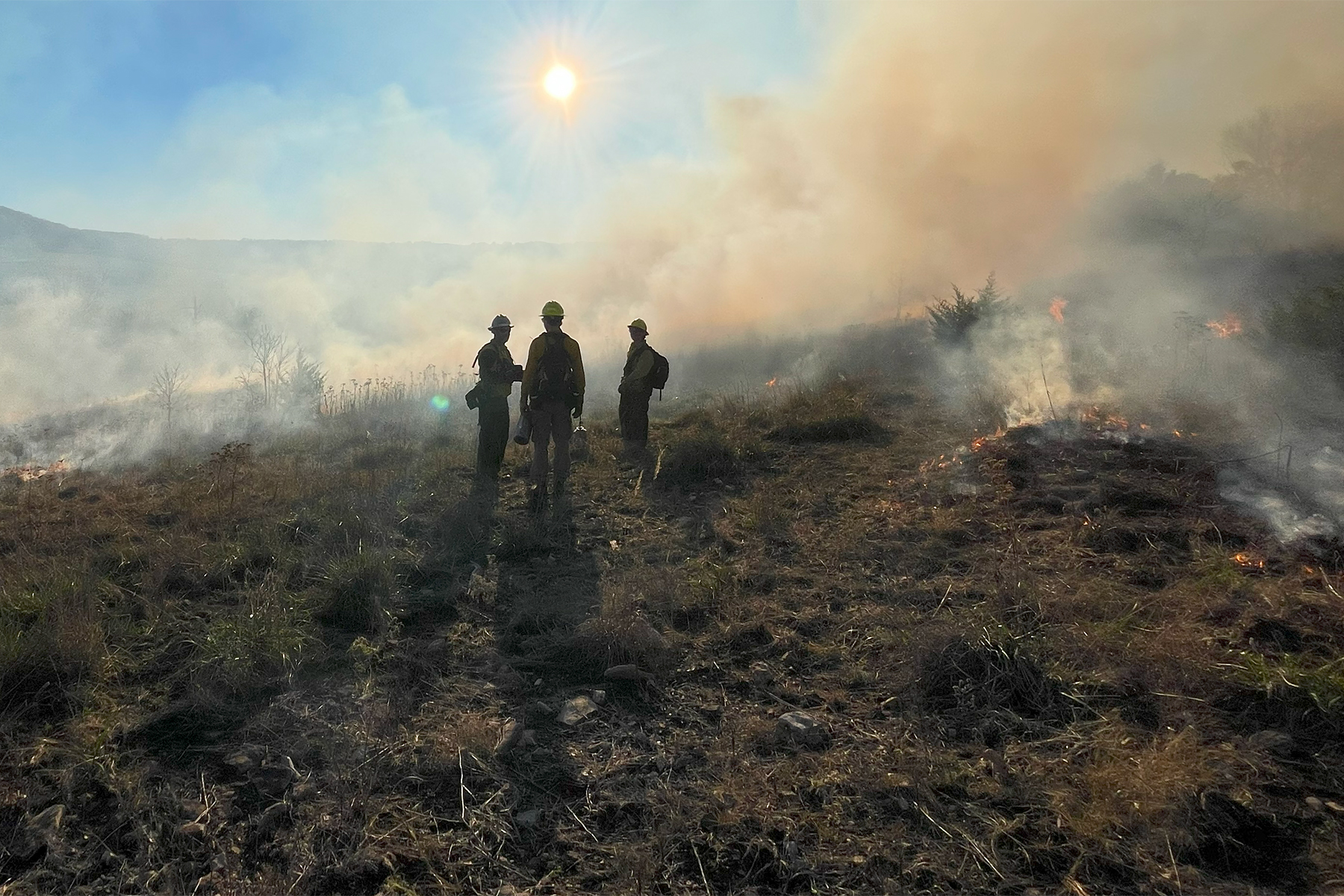 Three people silhouetted against smoke with burned ground all around them and a bright sun in the blue sky above them.