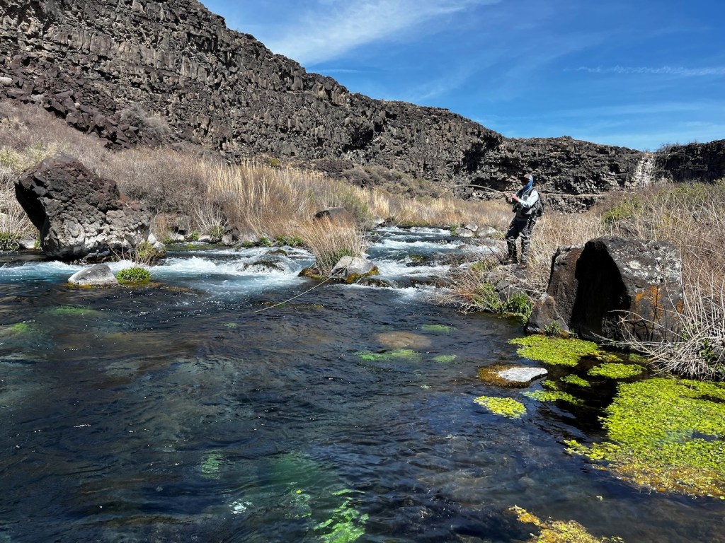 A man fishing at the base of a large basalt cliff. A stream with clear water is visible in the foreground.