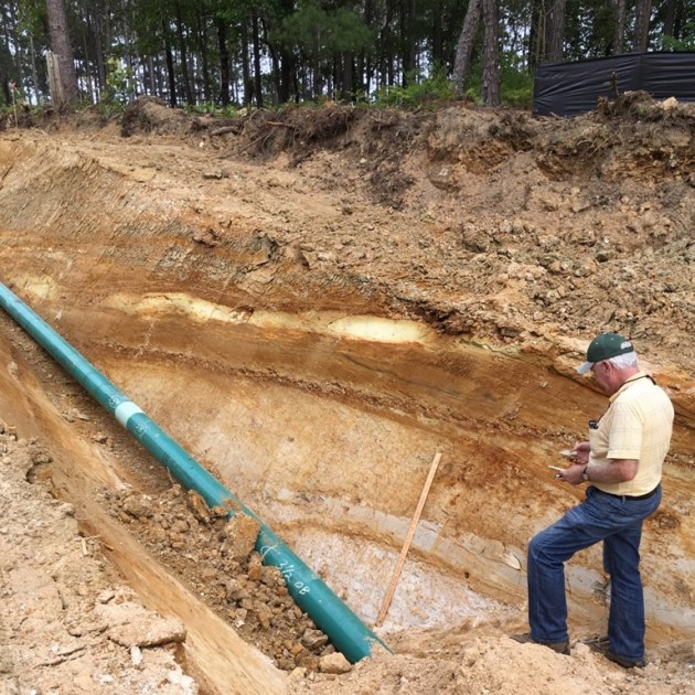 A man standing in a trench. The wall of the trench shows multicolored layers of sediment. 