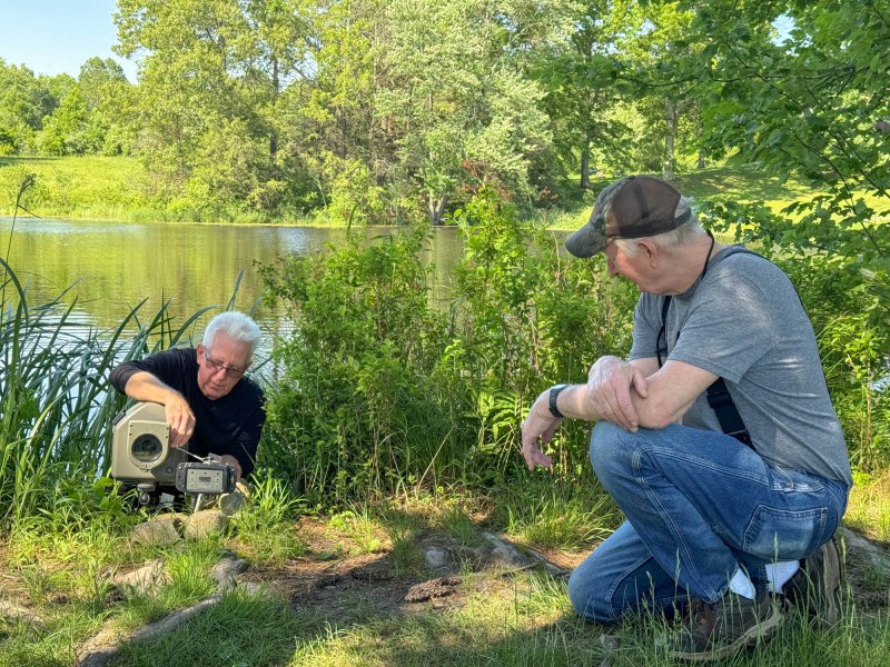 Two men set up a camera near a pond.