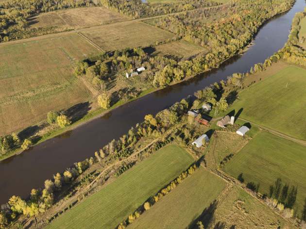 An aerial view of the Mississippi River flowing through a rural landscape. The river is bordered by dense trees on both sides, with patches of farmland and open fields extending beyond the tree line. Several farm buildings, including silos and barns, are visible near the riverbank. The fields are a mix of green and brown, indicating different stages of cultivation or crop types.