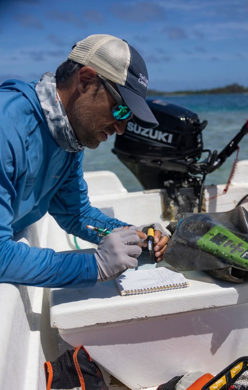 A man wearing fishing clothing, a hat and sunglasses takes notes on the back of a boat.