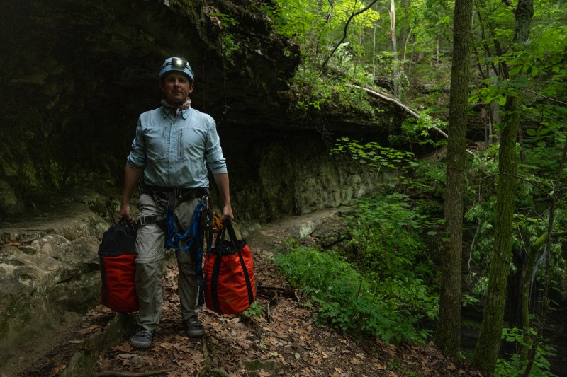 A man holding two bags of photo gear at the entrance to a cave.