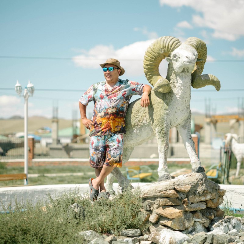 A man in a hat and sunglasses leaning against the statue of a large ram. 