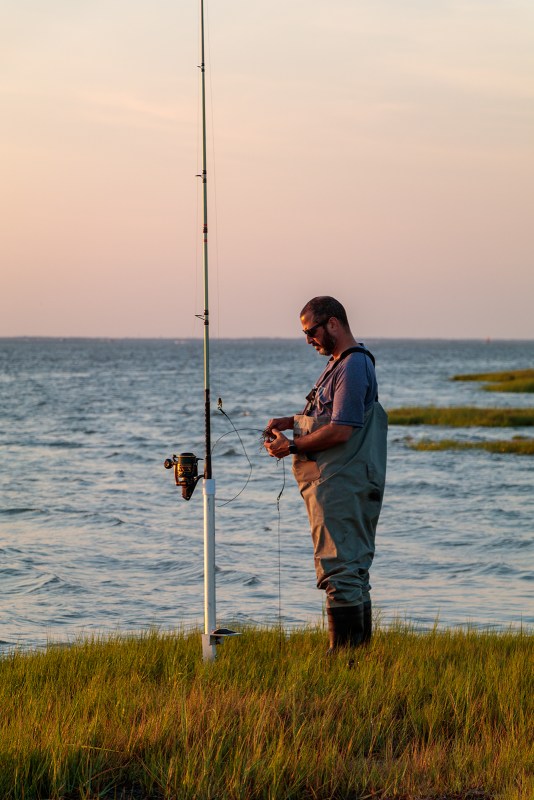Man in waders working with a fishing rod at sunset on the edge of a marsh with water in the background