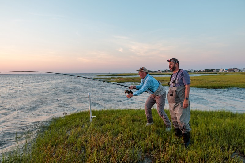 Two men in a salt marsh while one reels a fishing pole.