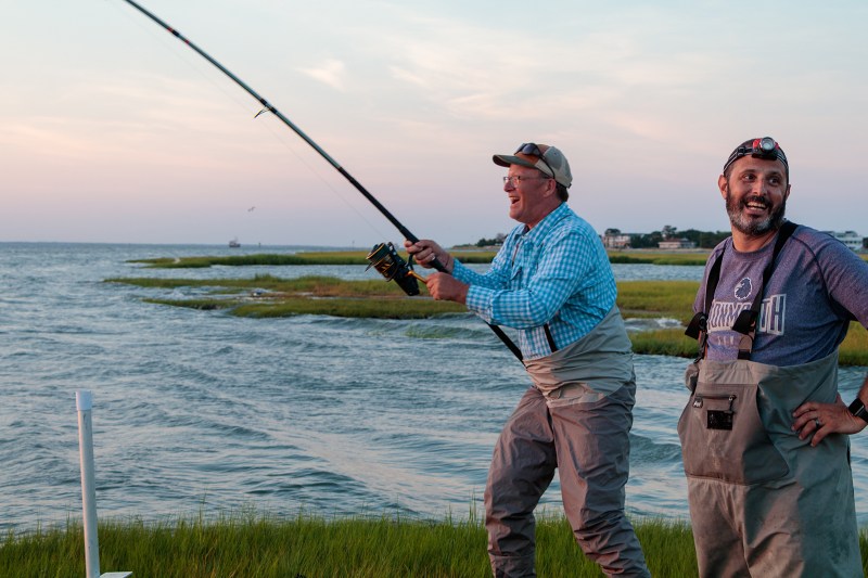 Two men standing on the edge of a marsh, one reeling in a fish on a bent fishing rod