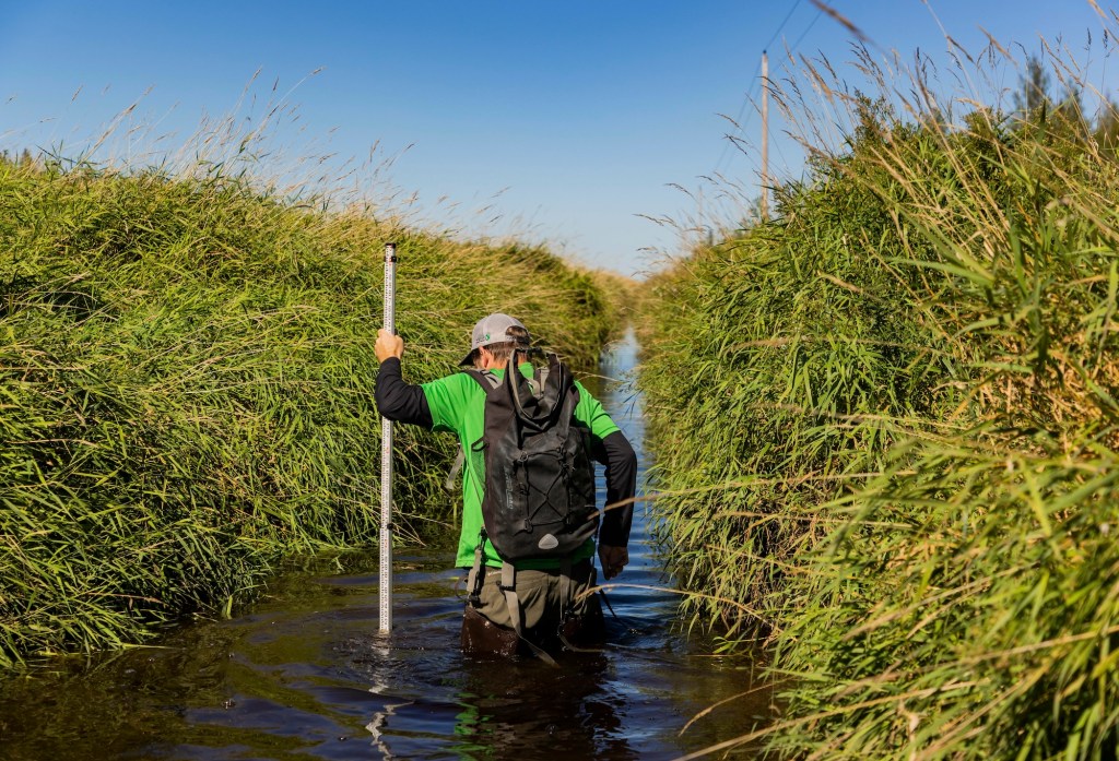 A man wearing a backpack walks through thigh-high water.