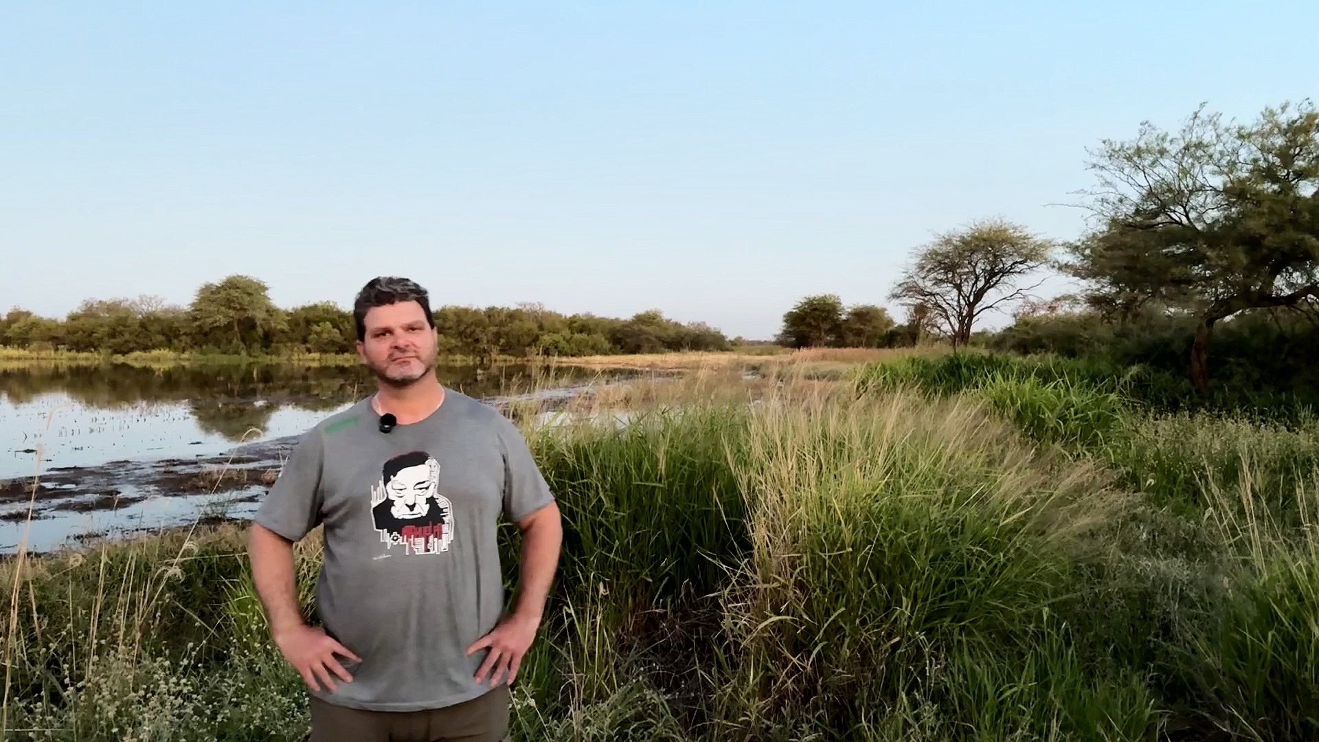 Man standing in front of small body of water, trees and tall grasses in the background.